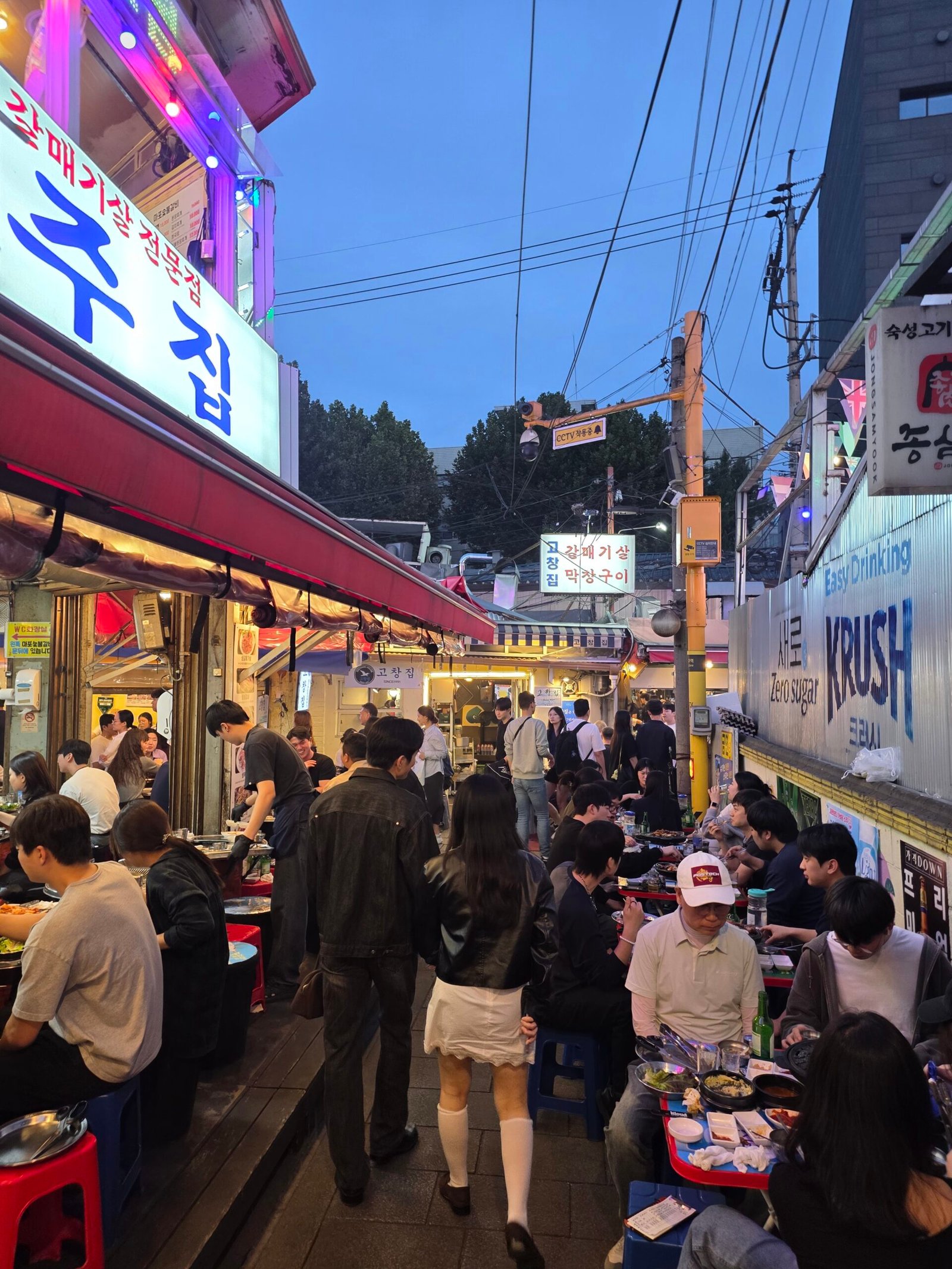 People moving between lit restaurants and outdoor tables in a Jongno 3-ga alley.
