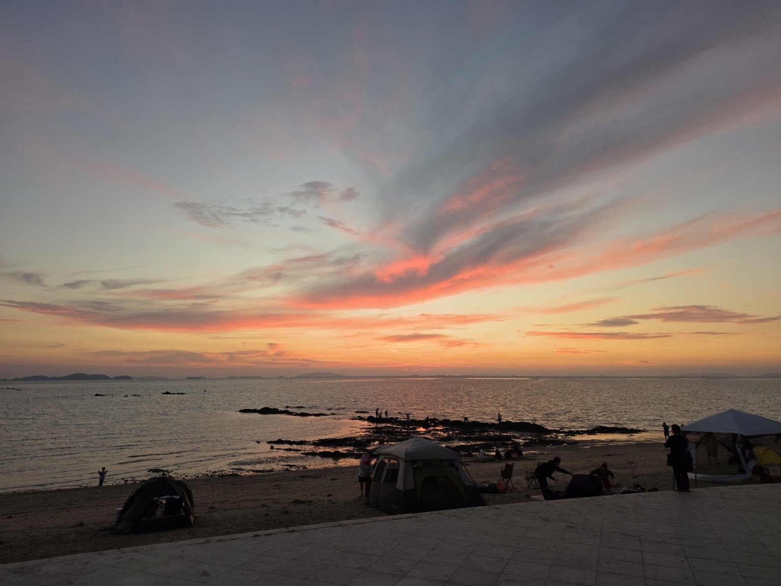 Pink and orange evening sky above Jebudo beach, with exposed rocks and small tents near the shore